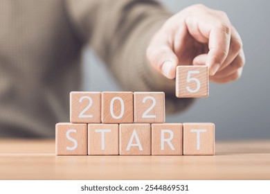 A businessman arranges wooden blocks spelling out 'START 2025' on a table, symbolizing the beginning of new goals and ventures for the upcoming year. Image reflects motivation and forward planning - Powered by Shutterstock