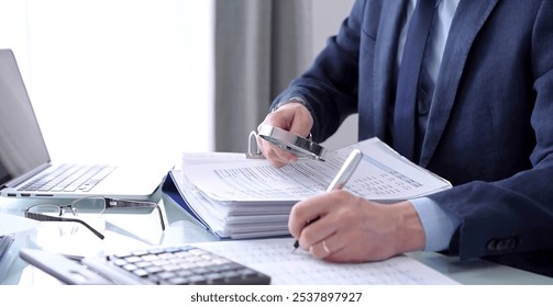 Businessman or auditor is writing notes while using a calculator and magnifying glass while financial documents analyzing. Audit and taxes concept