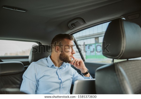 A businessman with a\
beard, sitting in the back seat of a car. He looks thoughtfully out\
the window