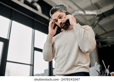 A businessman with a beard stands in a modern office, looking stressed as he takes an urgent phone call. - Powered by Shutterstock