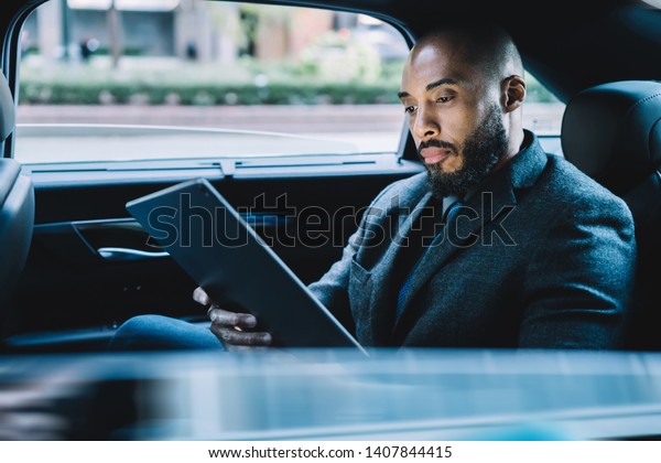 Businessman with black skin sitting on backseat in\
taxi automobile and searching contact number for making online\
video conference via modern touch pad, concept of market\
digitalisation on\
tablet