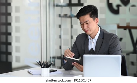 Businessman In Black Suit Working With Laptop Computer And Making Note In Notebook While Sitting At Office Desk.