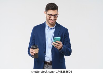 Businessman in blazer and eyeglasses, holding smartphone in one hand and takeaway coffee cup in another, looking at screen of phone, isolated on gray background - Powered by Shutterstock