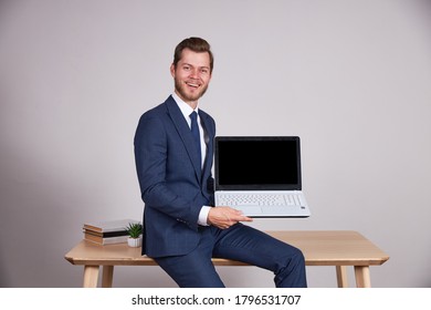 A Businessman In A Blue Business Suit Sits On A Wooden Office Table And Holds A Laptop, Look At The Camera And Smile And Show A Black Laptop Screen. White Background.