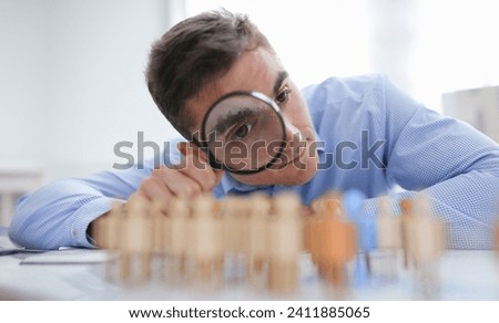 A businessman in blue shirt is holding a magnifying glass in his hand, is searching for personnel or people. Detective looking for missing person crowd of miniature figures choosing most suitable one