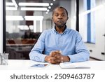 Businessman in blue shirt sits at office desk with hands clasped, conveying confidence and focus. Modern office environment with open book and phone, highlighting a professional work setting.