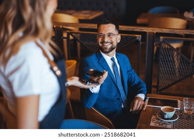 A businessman in a blue suit smiles while interacting with a waitress in a cozy cafe. The scene conveys a professional yet relaxed atmosphere with friendly service. - Powered by Shutterstock