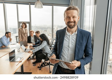 A businessman in a blue suit stands in a modern office, holding a tablet and smiling confidently. He looks directly at the camera. Several other people in business attire are sitting at a table - Powered by Shutterstock