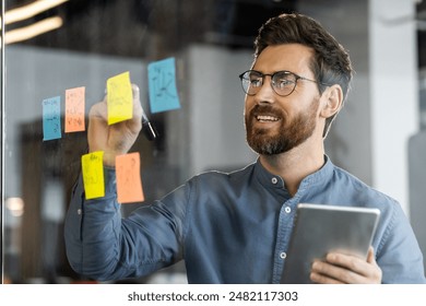 Businessman brainstorming and planning ideas on glass board using sticky notes while holding a tablet. Engaged in creative process and teamwork in modern office environment - Powered by Shutterstock