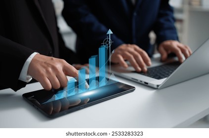 A businessman and businesswoman collaborate over a laptop, while the businesswoman points at a tablet displaying a holographic growth chart. - Powered by Shutterstock