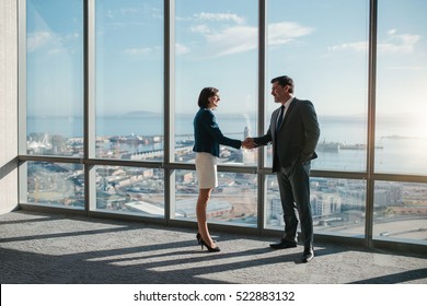 Businessman and businesswoman shaking hands together while standing in front of office building windows overlooking the city