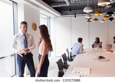 Businessman And Businesswoman Standing In Modern Boardroom Having Informal Discussion With Colleagues Meeting Around Table In Background