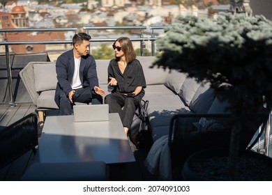 Businessman and businesswoman talking on balcony on building roof. Caucasian woman and asian man sitting on sofa at desk with laptop. Concept of business cooperation. Blurred background of city - Powered by Shutterstock