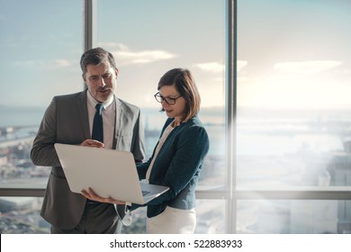 Businessman and businesswoman using a laptop together while standing in front of office building windows overlooking the city - Powered by Shutterstock