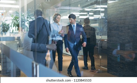 Businessman and Businesswoman Walking Through Glass Hallway, Discussing Work and Using Tablet Computer. Busy Corporate Office Building with Many Workers.