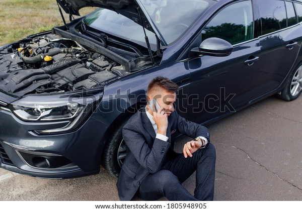 Businessman Calling for\
Roadside Assistance. Upset young man use phone sitting on road near\
the broken car opened the hood help repair stress problem emergency\
insurance auto