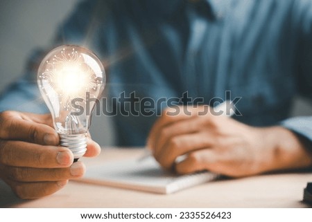 Businessman captures a good idea, holding a light bulb in his left hand. Symbol of solution and success. Working in an inspiring office, the author finds inspiration with a pen on paper.