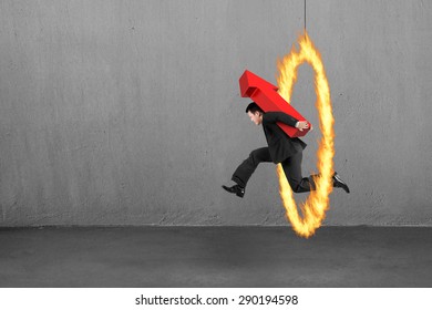 Businessman carrying 3D red arrow up sign, jumping through fire hoop, with concrete wall and floor indoors background.