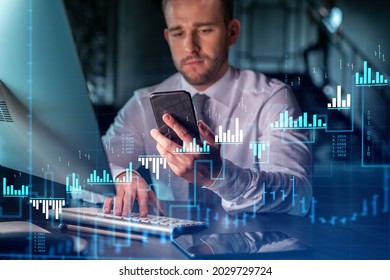 Businessman In Casual Wear Using Smart Phone To Optimize Trading Strategy At Corporate Finance Fund. Forex Chart Hologram Over Modern Office Background At Night Time