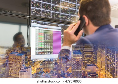 Businessman With Cell Phone Trading Stocks. Stock Analyst Looking At Graphs, Indexes And Numbers On Multiple Computer Screens. New Your City Lights Reflection In Glass.