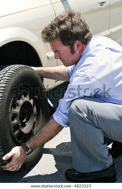 A businessman changing a flat tire on the road, red\
faced from the heat.