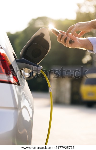 Businessman charging electric car at outdoor\
charging station Unrecognizable man unplugging electric car from\
charging station. Male unplugging in power cord to electric car\
using app on\
smartphone.