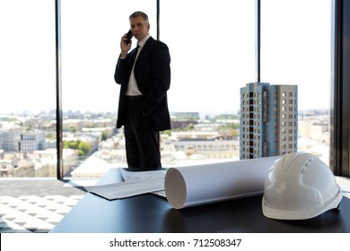 Businessman In Construction Company Office, Model Of Modern Multi Storey Residential Building House, Hardhat, Blueprint And Documents On Table