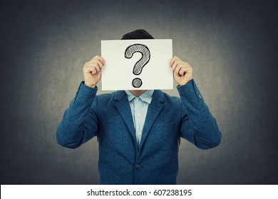 Businessman covering his face using a white paper with drawn question mark, like a mask, for hiding his identity. Isolated gray wall background.