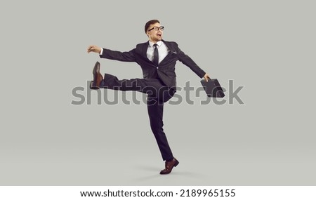 Businessman with crazy facial expression walking strangely, isolated on gray background. Full length of young man in suit with briefcase behaves strangely, takes wide step and shouts something.