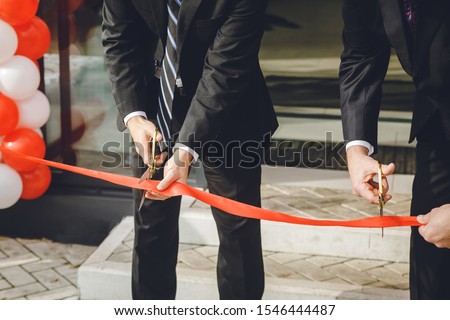 Businessman cutting red ribbon with pair of scissors. Two man in a classic black official suit cuts a red tape opening place