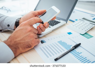 Businessman At Desk Using A Financial App On His Smart Phone And Working On Reports, Hands Close Up