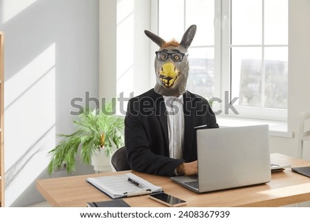 Businessman in donkey head mask using laptop computer in office. Funny stupid male office employee in suit, rubber mask and glasses sitting at desk with laptop and looking at camera