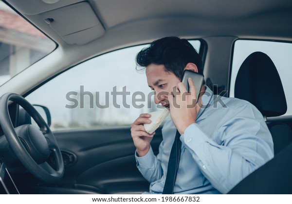Businessman eating sandwich while working in\
laptop and talking on the phone  in the drivers seat in his car.\
Busy businessman and food in\
car.\
