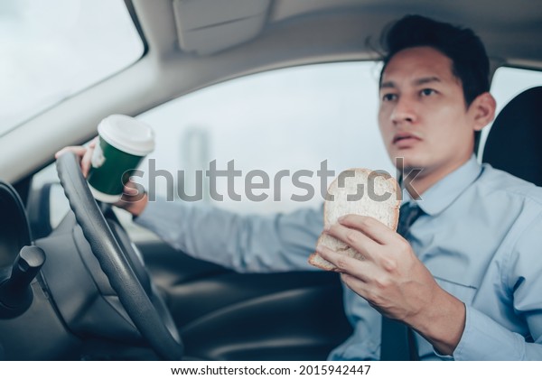 A\
businessman eating whole wheat toast and holding a coffee cup While\
driving on the road and he did not lose a seat\
belt.