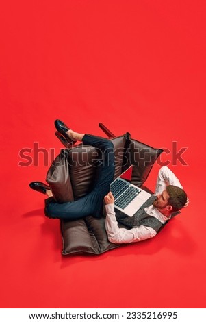 Businessman, employee sitting on armchair in weird, uncomfortable position, working on laptop against red studio background. Concept of business, working routine, deadlines, freelance, office, ad