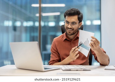 Businessman engaging in video call holding growth chart, analyzing data at office. Professional sitting at desk with laptop, focusing on presentation and communication in contemporary workplace. - Powered by Shutterstock