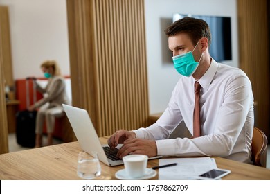 Businessman With Face Mask Using Computer And Typing An E-mail While Working In Hotel Room. His Female Colleague Is In The Background. 