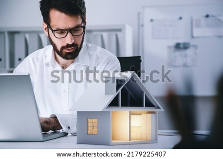 Businessman in formal wear is typing on laptop looking at model of paper house. Real estate insurance agency. Office in background. Concept of financial protection, indemnity, modelling, styling