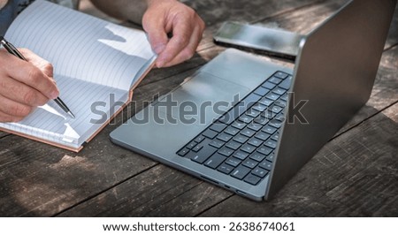 Businessman or freelancer writing in notebook while working outdoors on laptop at wooden table. Creative remote work or planning concept