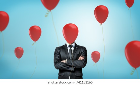 A businessman in front view with crossed arms stands surrounded by many red party balloons with one hiding his face. Businessman in disguise. Entertainment business. Birthday at work.