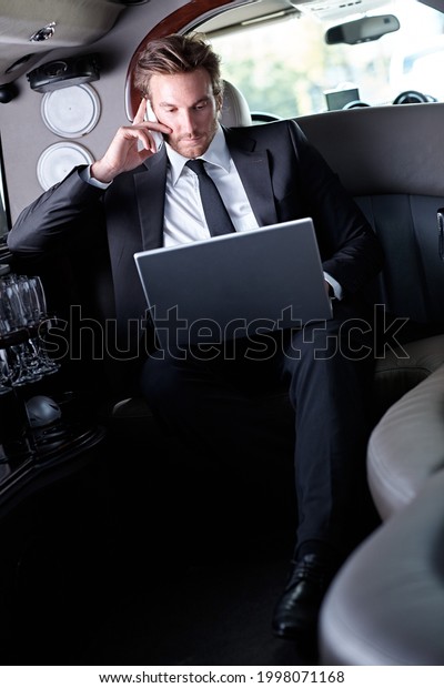 Businessman in full suit traveling in luxury\
limousine. Sitting in car working on laptop calling on phone.\
Business travel\
concept.