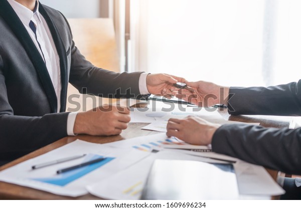 businessman giving car key to his colleague,\
and reaching over receiving the car key, with work on paper about\
statistics and data about the business, piling on top of the table\
with pen and\
clipboard