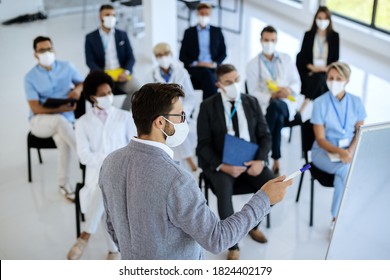Businessman giving a presentation and wearing protective face mask while talking to group of entrepreneurs and healthcare workers in convention center.