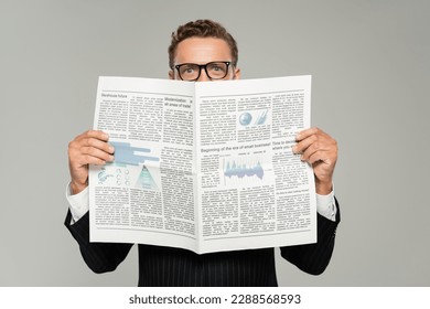 businessman in glasses looking at camera while covering face with newspaper isolated on grey