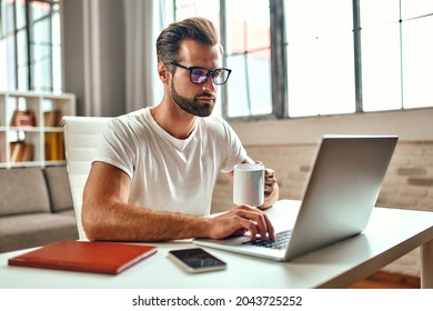 Businessman in glasses with a mug of coffee works at a laptop. Freelance, work from home. - Shutterstock ID 2043725252