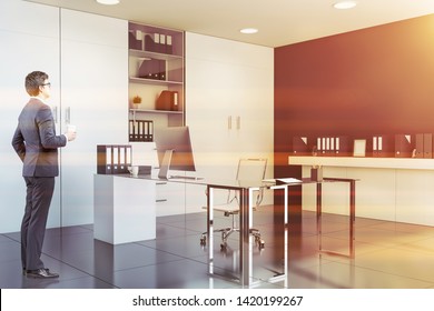 Businessman In Glasses Standing In CEO Office With Black Walls, White Bookcase And Glass Computer Table. Concept Of Manager Lifestyle. Toned Image