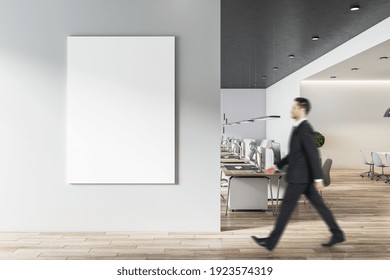 Businessman Goes To Blank White Poster On Light Grey Wall In Modern Open Space Office With Light Furniture And Wooden Floor. Mock Up