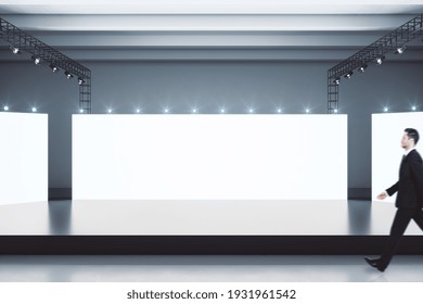 Businessman goes through black stage in spacious showroom with blank white glowing screens behind. Mockup