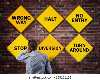 Businessman Going The Wrong Way In Front Of A Brick Wall With Stop, Wrong Way, Halt, No Entry, Diversion And Turn Around Road Signs Concept For Blocked Direction In Business