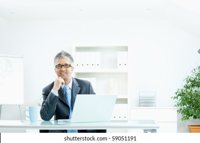 Businessman With Grey Hair, Wearing Grey Suit And Glasses Thinking Over Laptop Computer, Sitting At Desk In Bright, Modern Office, Leaning On Hand, Smiling.